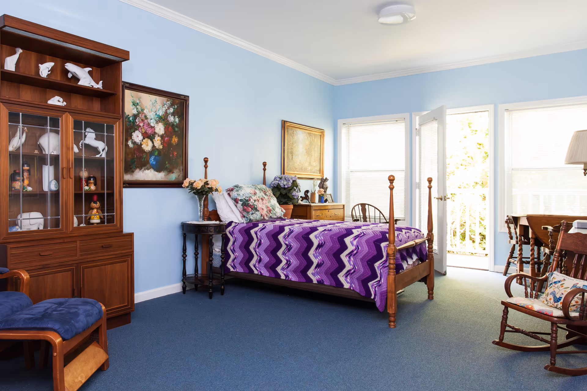 A cozy bedroom with light blue walls and blue carpet featuring a wooden four-poster bed covered with a purple zigzag patterned blanket and floral pillows. There is a wooden cabinet with glass doors displaying white animal figurines and other decorative items. A small round side table with a vase of flowers is next to the bed. The room also includes a wooden rocking chair with a floral cushion, a wooden dresser with plants and framed pictures, and a door leading to a balcony with windows letting in natural light.