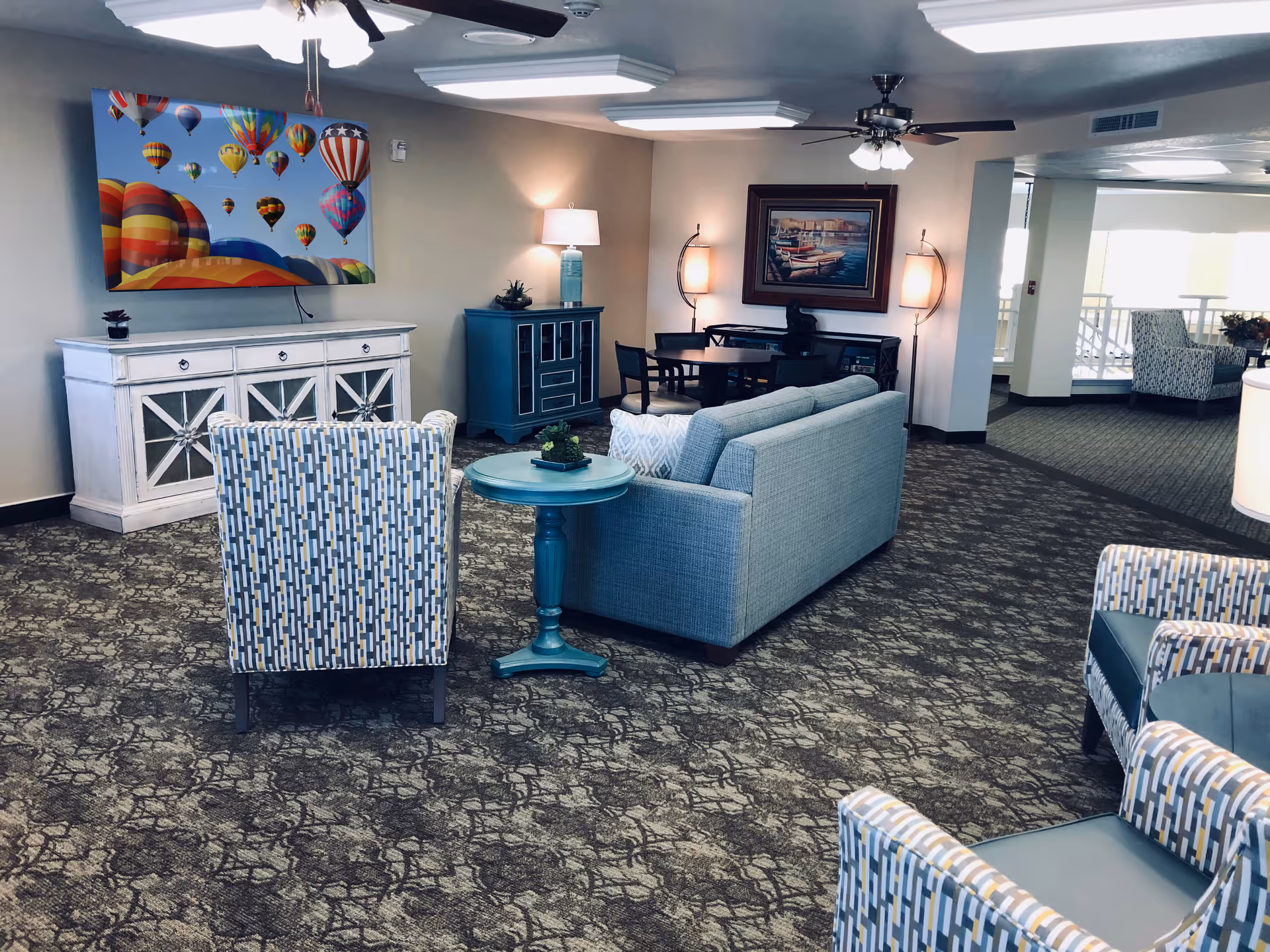 A spacious and well-lit living room area in a senior living facility featuring a light blue sofa, patterned armchairs, a round blue side table, and a white cabinet with a colorful hot air balloon painting above it. The room has carpeted flooring, ceiling fans with lights, and additional seating and tables in the background near large windows.