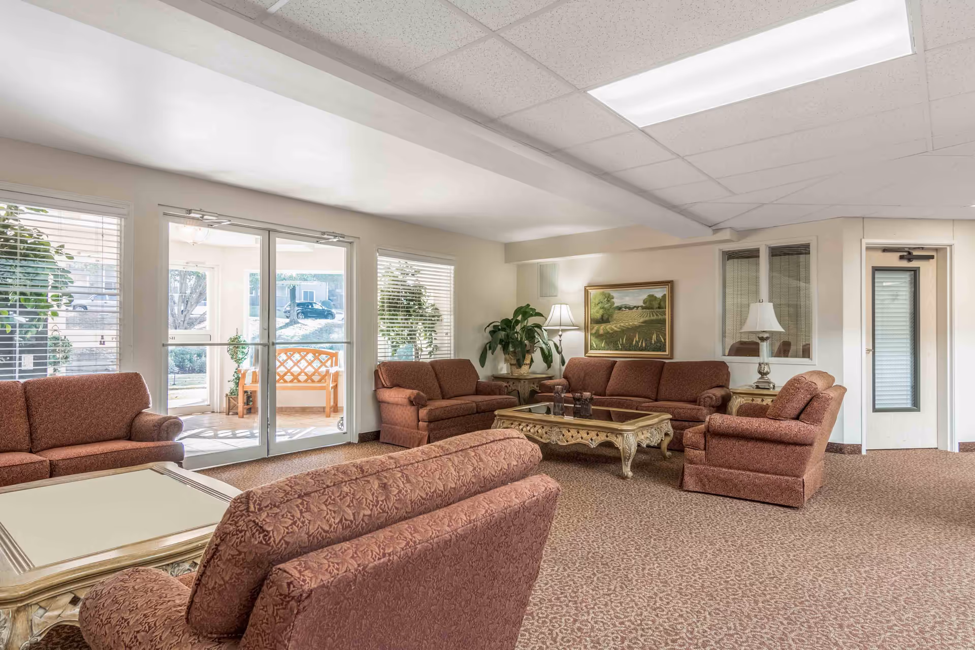 A spacious living room area with multiple upholstered sofas and armchairs in a reddish pattern, a decorative coffee table, two side tables with lamps, a large plant, and a framed landscape painting on the wall. Large windows and glass doors let in natural light and show an outdoor bench and greenery.