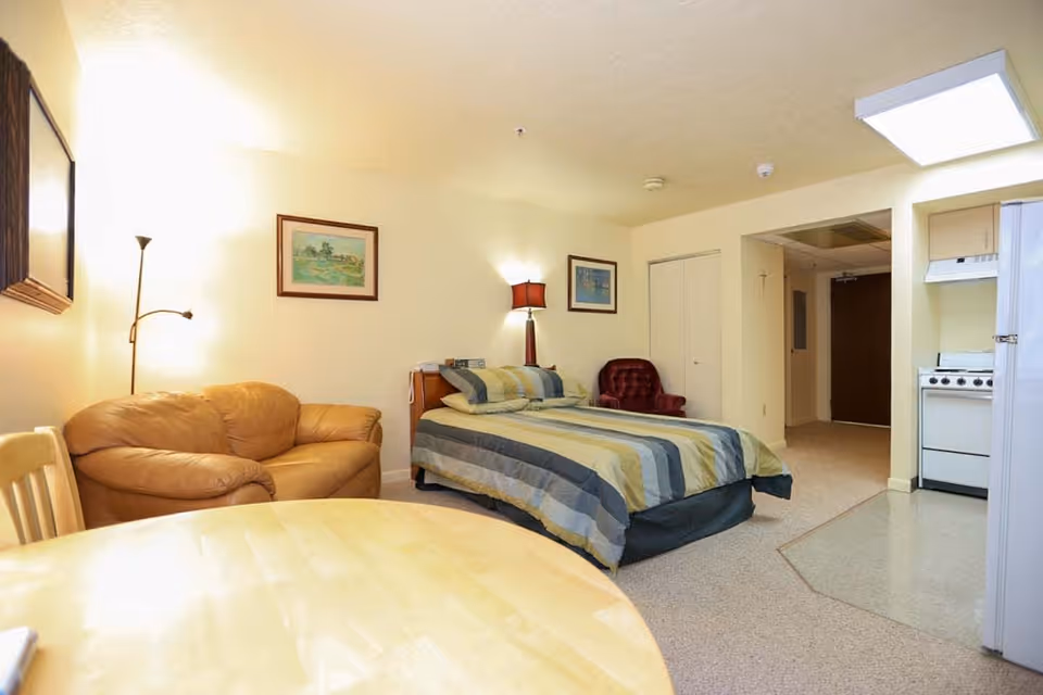Interior view of a senior living facility room featuring a bed with striped bedding, a brown leather loveseat, a red armchair, a wooden table with chairs, and a small kitchen area with a stove and refrigerator. The room is warmly lit with lamps and has framed artwork on the walls.