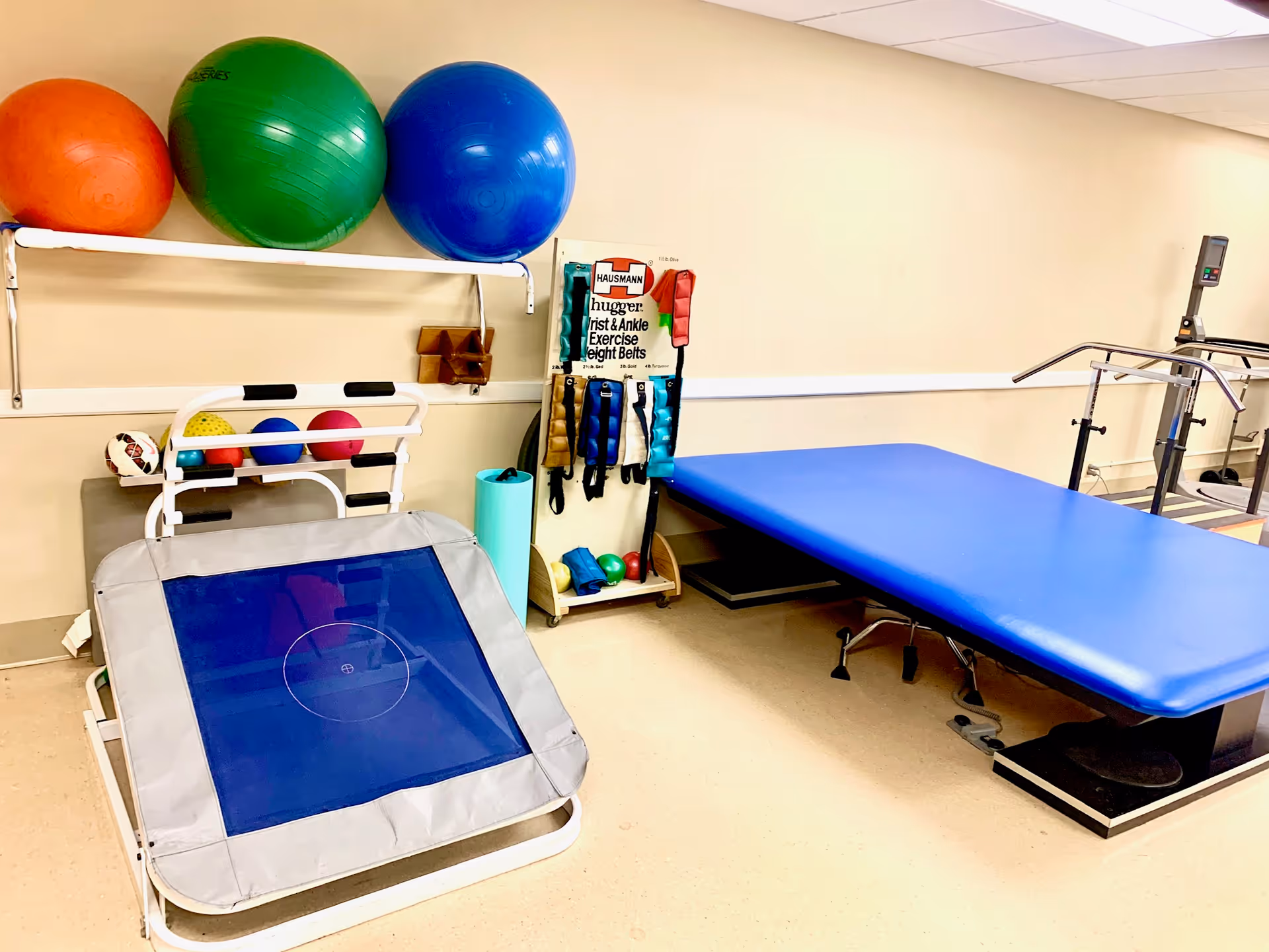 Physical therapy room with exercise equipment including a blue therapy table, a small trampoline, various exercise balls in orange, green, and blue, wrist and ankle exercise weight belts hanging on a rack, and a treadmill in the background.