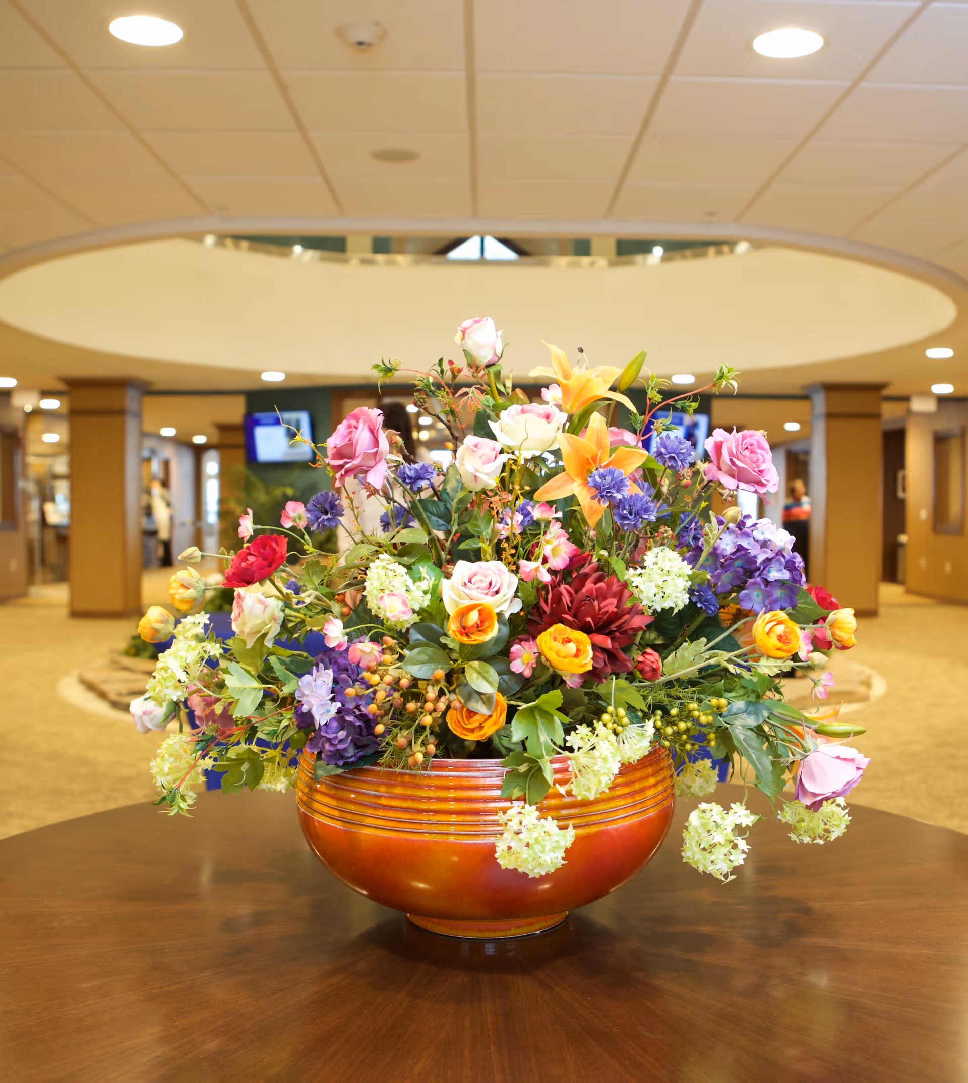 A large, colorful floral arrangement in an orange ceramic bowl sits on a wooden table in the center of a spacious, well-lit lobby area with beige carpeting and columns.