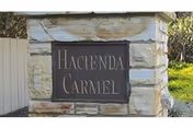 Stone entrance monument sign set in a stacked stone pillar reading "Hacienda Carmel" with grass and a fence visible behind it.