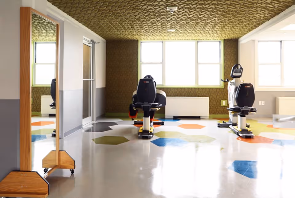 A bright exercise room with two recumbent stationary bikes facing large windows. The floor has colorful hexagonal patterns in green, blue, orange, and gray. The walls are painted gray and white with a textured green accent wall and ceiling. A full-length mirror on wheels is positioned on the left side of the room.