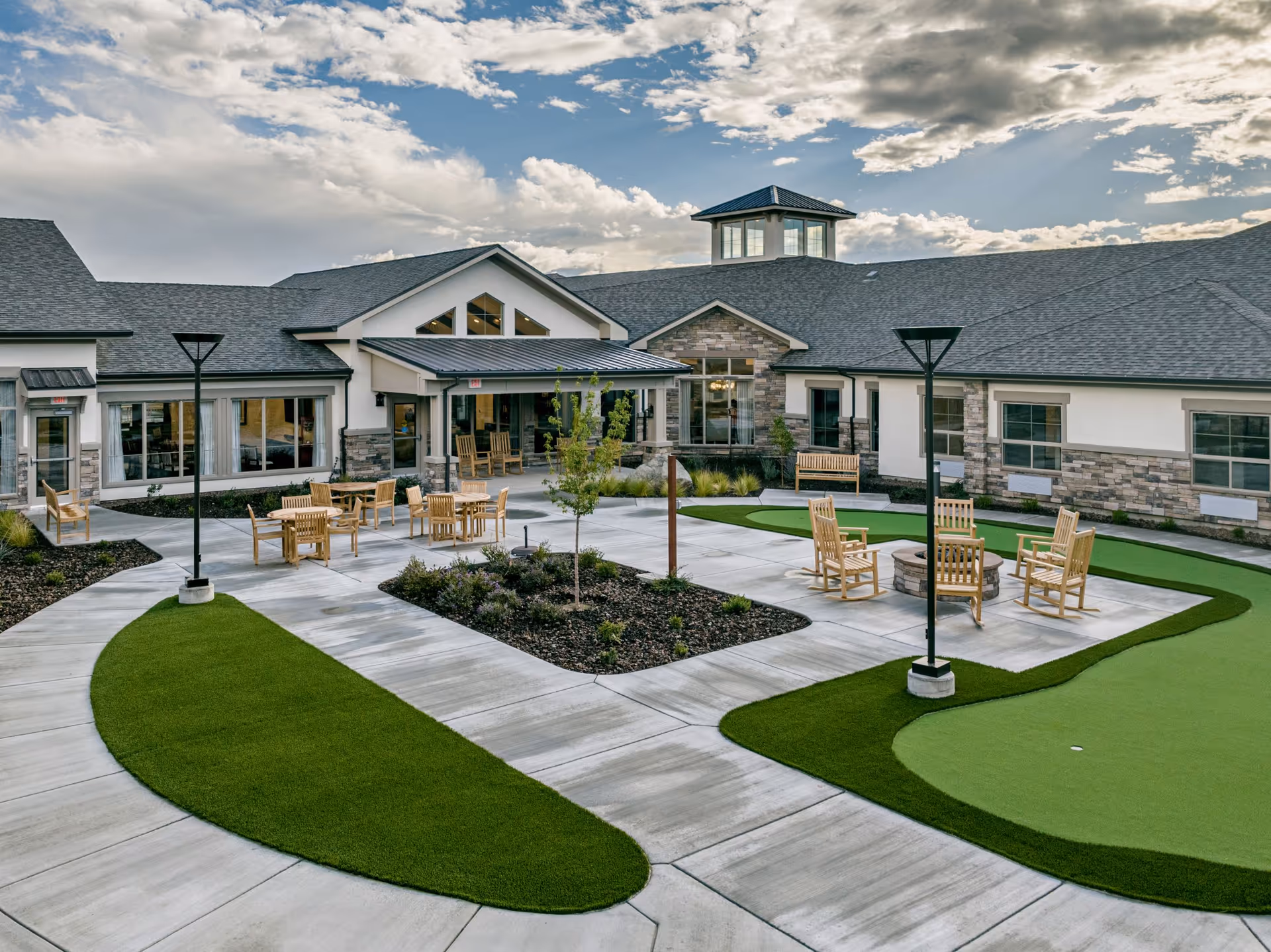 Outdoor courtyard area of Kiley Ranch Senior Living featuring a paved walkway, green artificial turf, wooden chairs arranged around a fire pit, tables with chairs, benches, and a building with large windows and a cupola under a partly cloudy sky.
