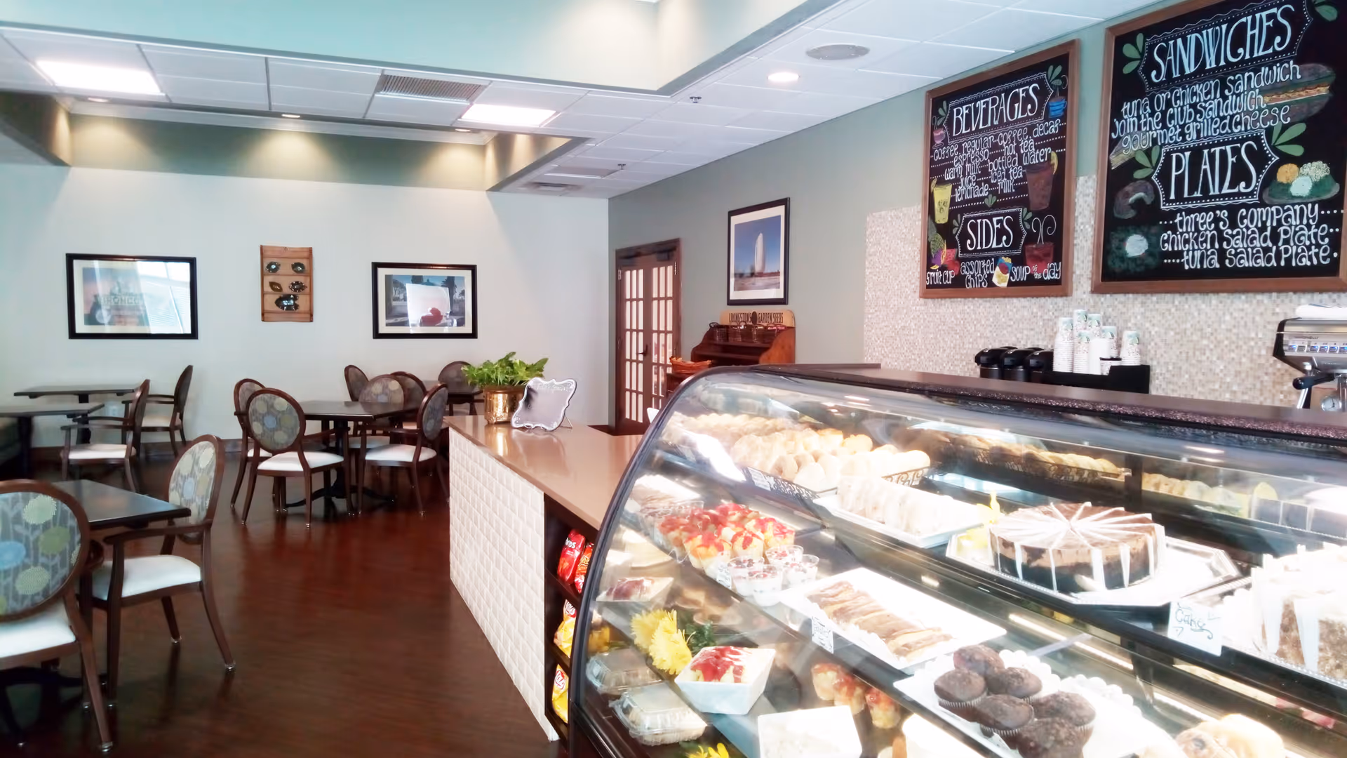 A brightly lit cafe-style dining area with a glass display case of pastries and desserts at the counter and several dining tables and chairs in the background.