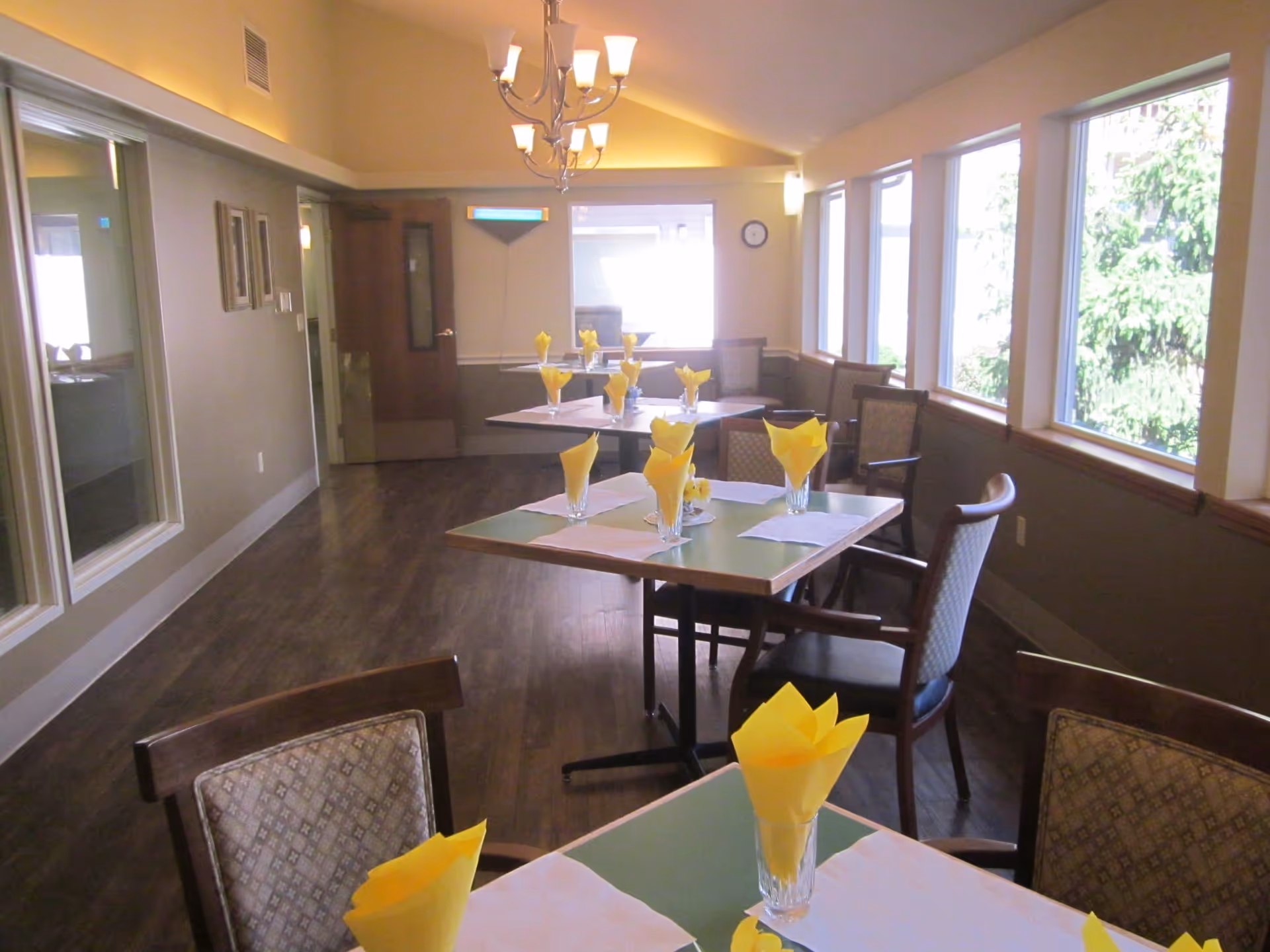 Dining room with several tables set with yellow folded napkins in glasses and white placemats. The room has large windows on one side letting in natural light, wooden chairs with patterned cushions, and a chandelier hanging from the ceiling.
