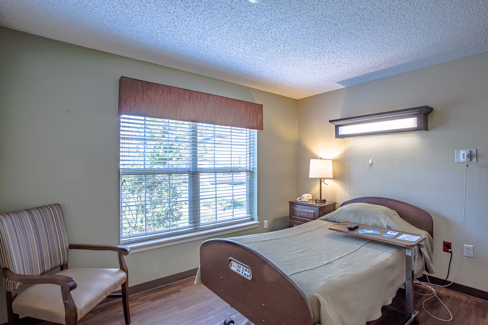 A simple senior living bedroom with a single bed covered in a beige blanket, a wooden side table with a lamp and telephone, a striped armchair, and a large window with blinds letting in natural light.