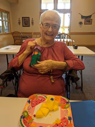 An elderly woman sitting in a wheelchair at a table in a well-lit room, smiling and holding a small colorful craft decoration. On the table in front of her is a plate with pieces of cut fruit, including pineapple. The room has wooden chairs and tables, with windows letting in natural light.