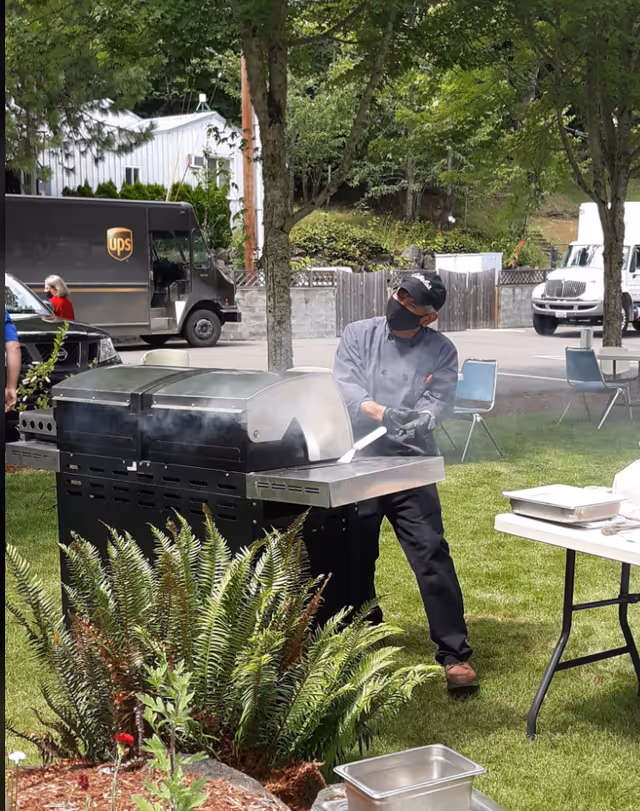 A person wearing a black face mask, black gloves, and a chef's uniform is grilling food on a large outdoor grill in a grassy area with trees and plants nearby. There are folding chairs and tables set up around, and a UPS delivery truck and other vehicles are visible in the background.
