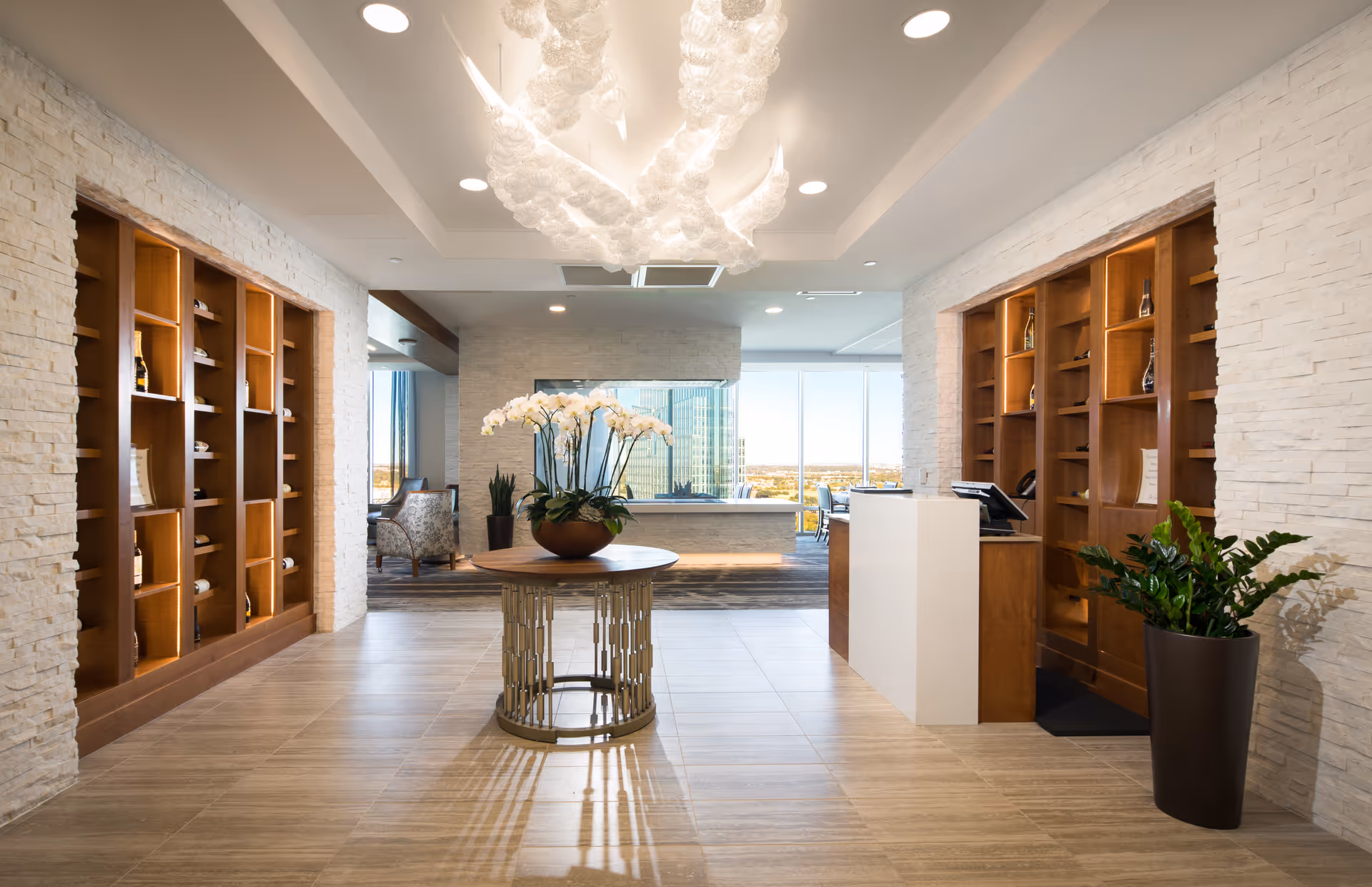 A bright and modern interior space with a round table in the center holding a large pot of white orchids. The room features light-colored tiled floors, white textured walls, and wooden shelving units on both sides displaying decorative items. A white reception desk is on the right side, and large windows in the background offer a view of a cityscape. A unique, artistic chandelier hangs from the ceiling.