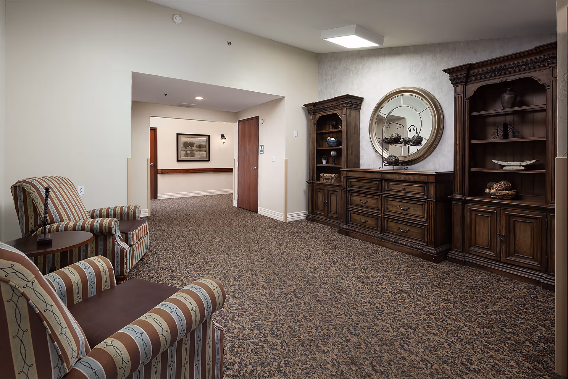 Carpeted common area with striped armchairs, a side table, and large wooden cabinetry topped by a round mirror in a senior living facility.