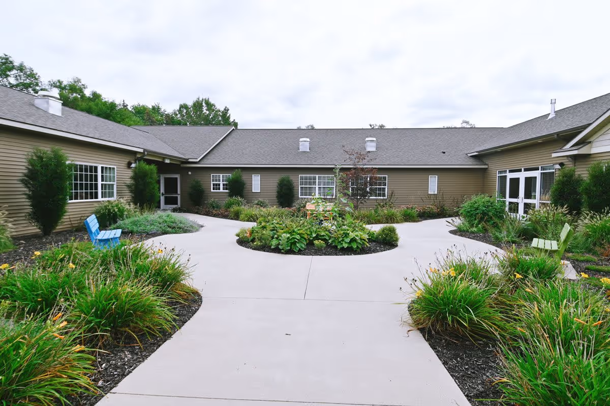 Outdoor courtyard area of The Cortland Wyoming facility featuring a concrete pathway that splits around a circular garden bed with various plants and a small tree in the center. The courtyard is surrounded by a single-story building with beige siding, multiple windows, and doors. There are green and blue benches placed along the pathway, and the area is landscaped with bushes and flowering plants under an overcast sky.