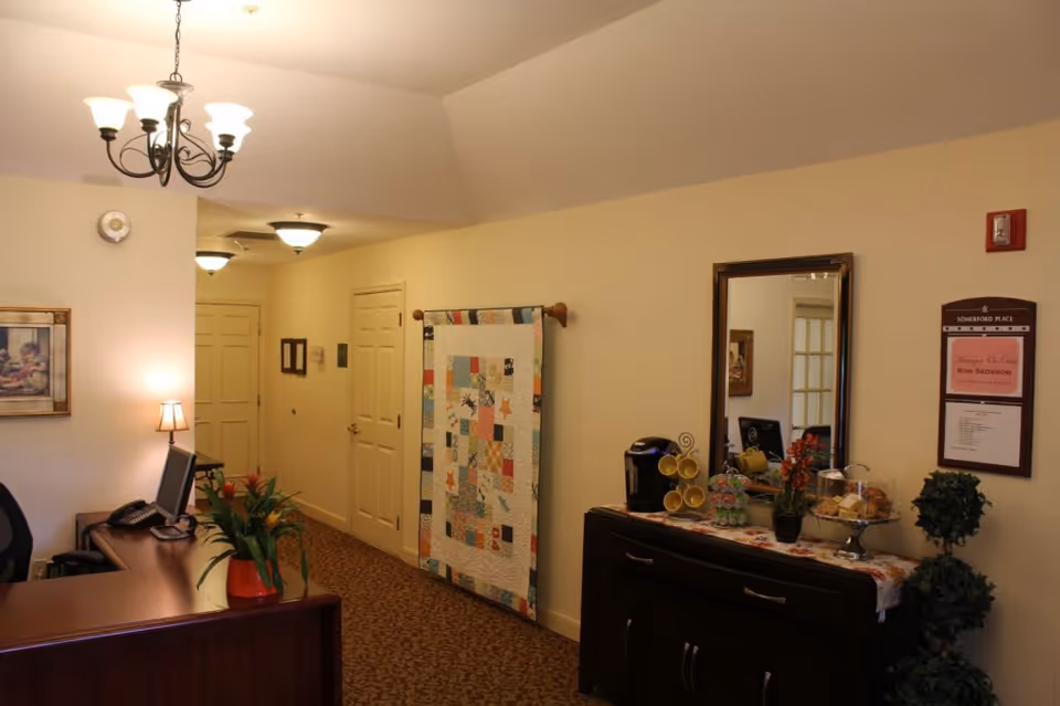Reception area inside a senior living facility with a desk, a quilt hanging on the wall, mirror, and buffet table with snacks and plants.