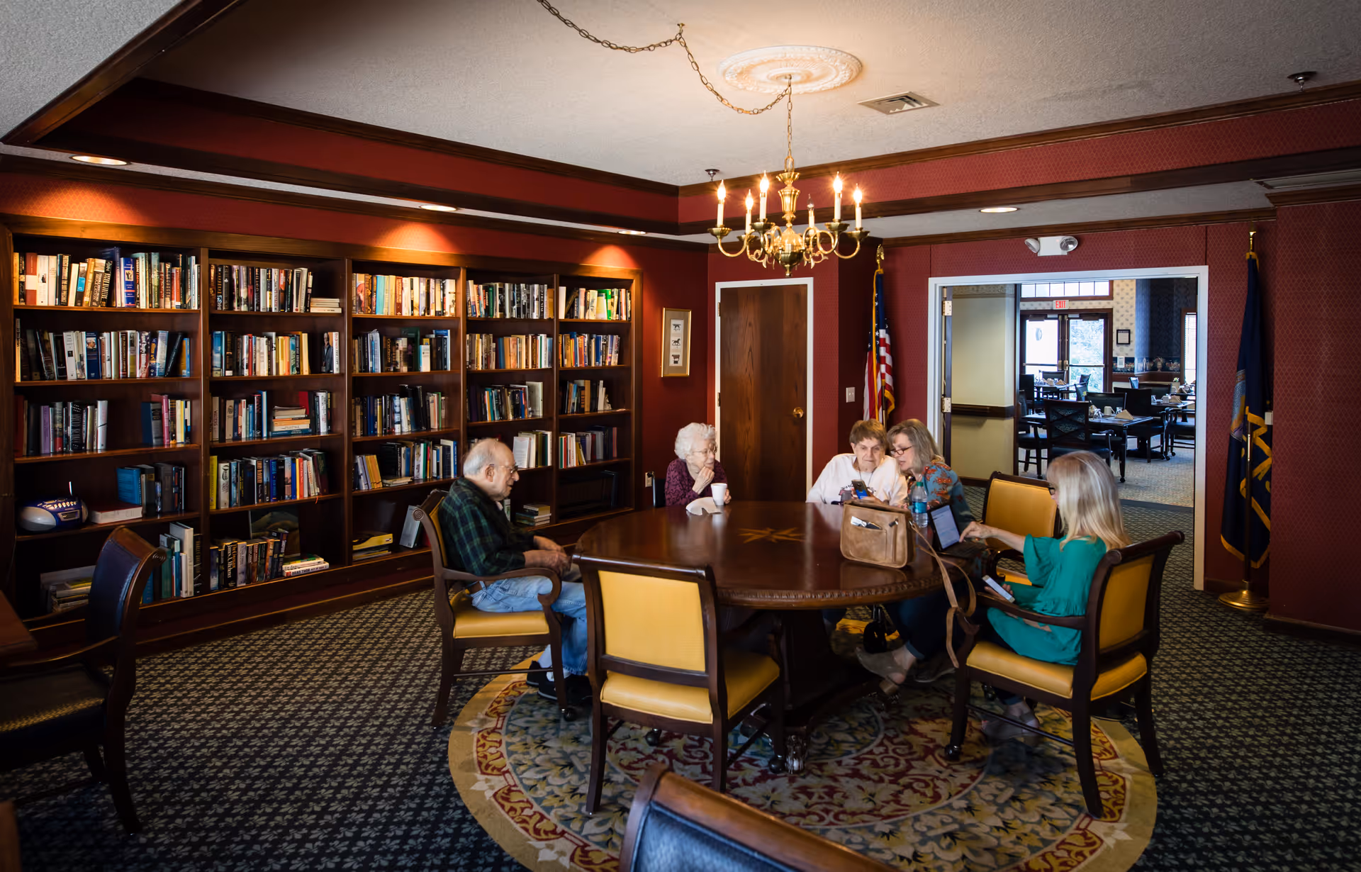 A cozy room with a large wooden round table surrounded by six chairs, five elderly people sitting and engaging in conversation or using a tablet. The room has a carpet with a decorative pattern, a chandelier hanging from the ceiling, and a large bookshelf filled with books along one wall. An American flag and another flag stand near a doorway leading to another room with more tables and chairs.