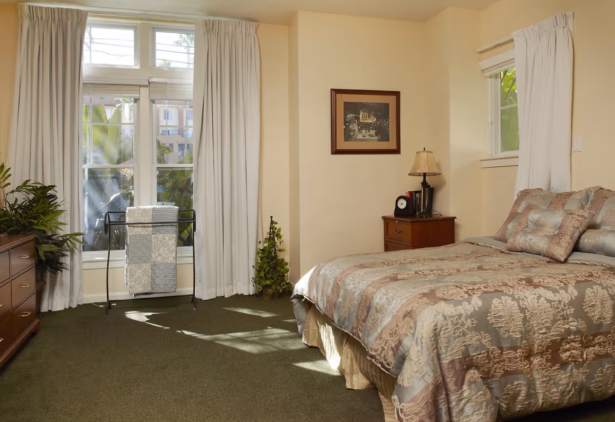 A cozy bedroom with a large bed covered in a patterned comforter and matching pillows. There is a wooden nightstand with a lamp, clock, and books. A framed picture hangs on the wall above the nightstand. The room has two windows with white curtains, a green carpet, a wooden dresser, and a plant stand with a folded quilt near the window.