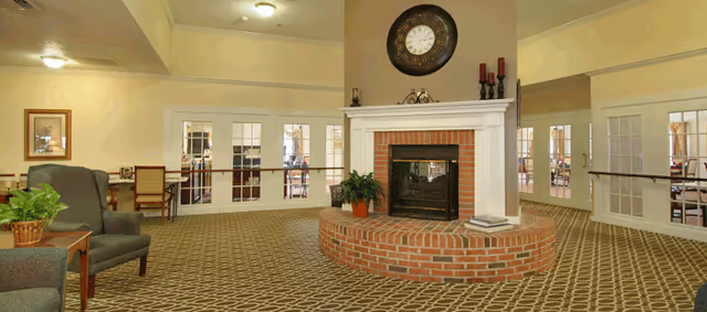 A spacious common area with a central brick fireplace topped by a large round clock. The room features patterned carpet, beige walls, and multiple glass-paneled doors leading to other rooms. There are chairs and a small table with a plant on the left side.