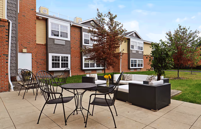 Outdoor patio with metal tables and chairs and wicker lounge seating on a paved area in front of a two-story brick senior living building with trees and lawn.