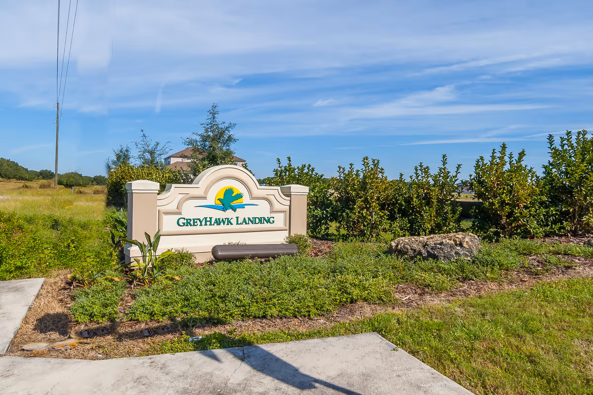 Outdoor view of a landscaped area with a sign that reads 'GreyHawk Landing' surrounded by greenery and bushes under a blue sky with some clouds.