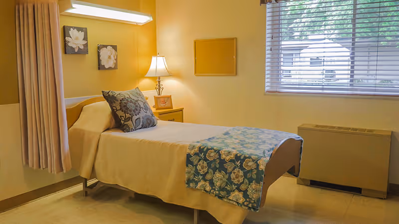 A single bed in a small, neatly arranged room with beige walls. The bed has a beige bedspread and a blue floral throw blanket at the foot, along with a decorative pillow. There is a wooden nightstand with a lamp and a framed picture next to the bed. Two floral paintings hang on the wall above the bed. A window with blinds lets in natural light, and a heating or air conditioning unit is below the window.