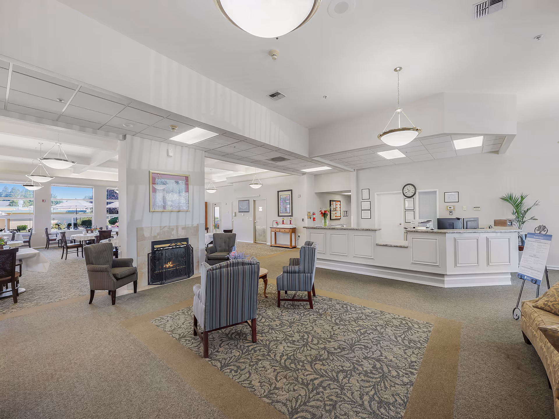 A spacious and well-lit assisted living facility common area featuring a reception desk on the right, a seating area with four upholstered chairs around a small table in the center, and a fireplace with a framed picture above it. In the background, there is a dining area with tables and chairs near large windows letting in natural light.