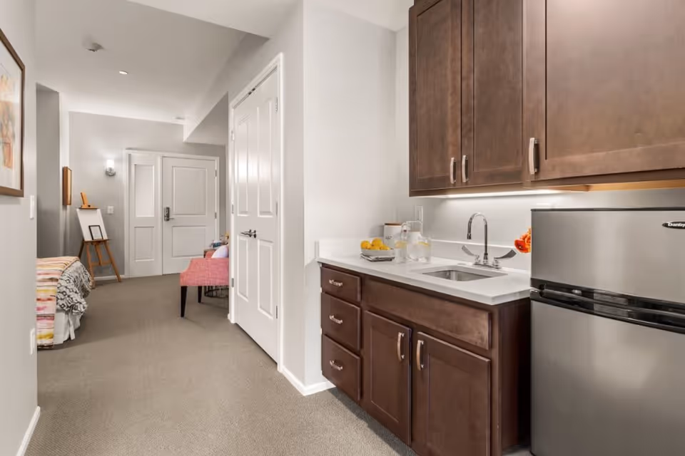 Interior view of a senior living facility room showing a small kitchenette with dark wood cabinets, a stainless steel mini refrigerator, a sink, and countertop with a glass pitcher and lemons. In the background, there is a hallway leading to a bed with a colorful blanket and a pink chair beside it. The walls are painted light gray and the floor is carpeted.