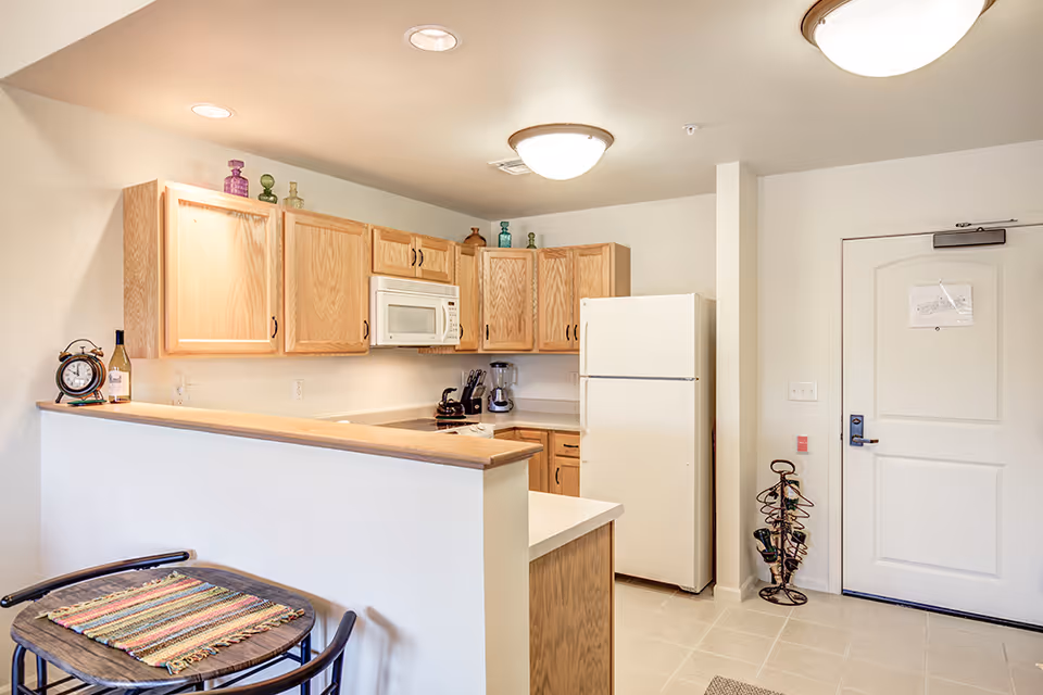 A compact kitchen area with light wood cabinets, a white refrigerator, a microwave, and a small countertop. There is a small table with two chairs and a colorful placemat in the foreground. The kitchen is well-lit with ceiling lights and has a white door to the right.