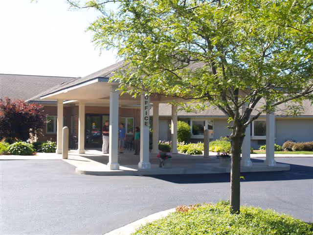 Exterior view of the entrance to Appledorn Assisted Living Center North, showing a covered drop-off area with columns, a tree in the foreground, and a few people standing near the entrance door.
