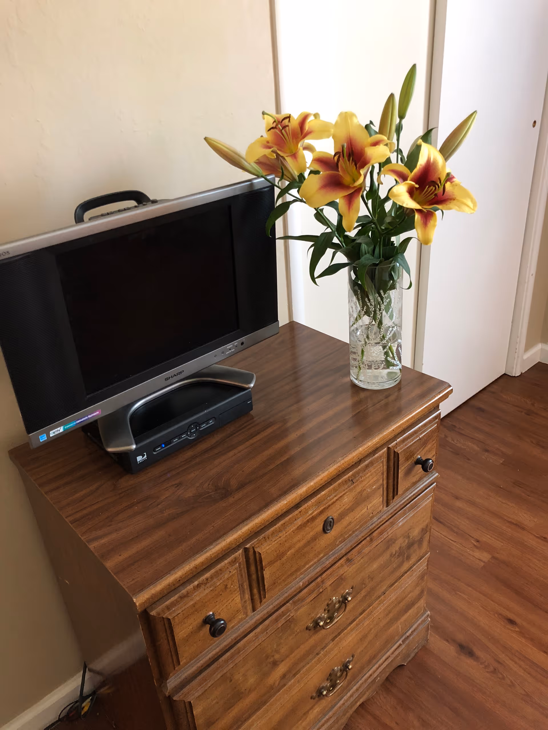 A wooden dresser with a small flat-screen TV and a glass vase holding yellow and red lilies on top. The dresser is placed against a beige wall and next to a white door, with wooden flooring visible.