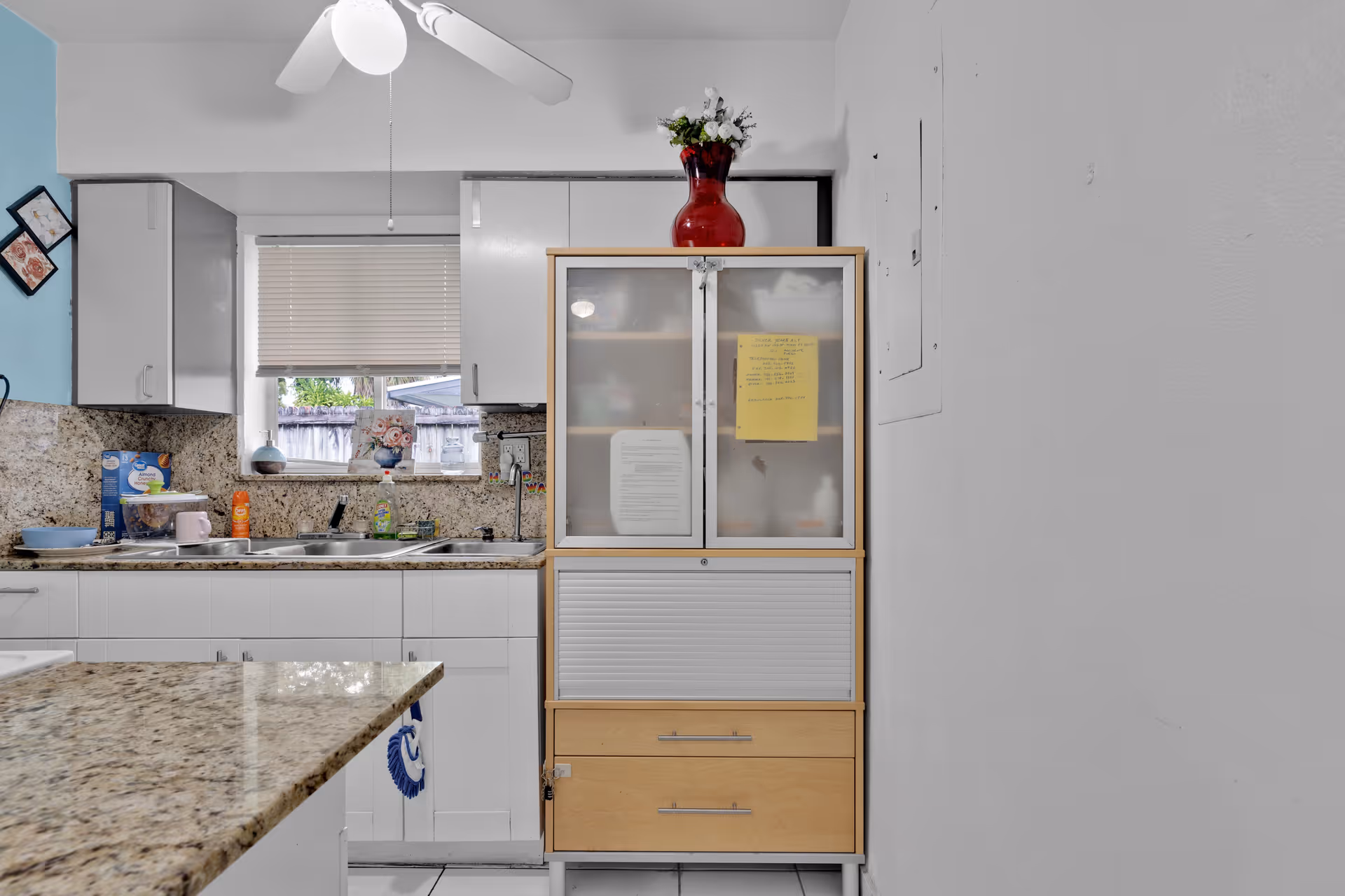 A kitchen interior featuring white cabinets, a granite countertop, a double sink under a window with closed blinds, and a wooden cabinet with frosted glass doors and drawers. A red vase with flowers is placed on top of the cabinet. The walls are painted white and light blue, and a ceiling fan with a light is visible.