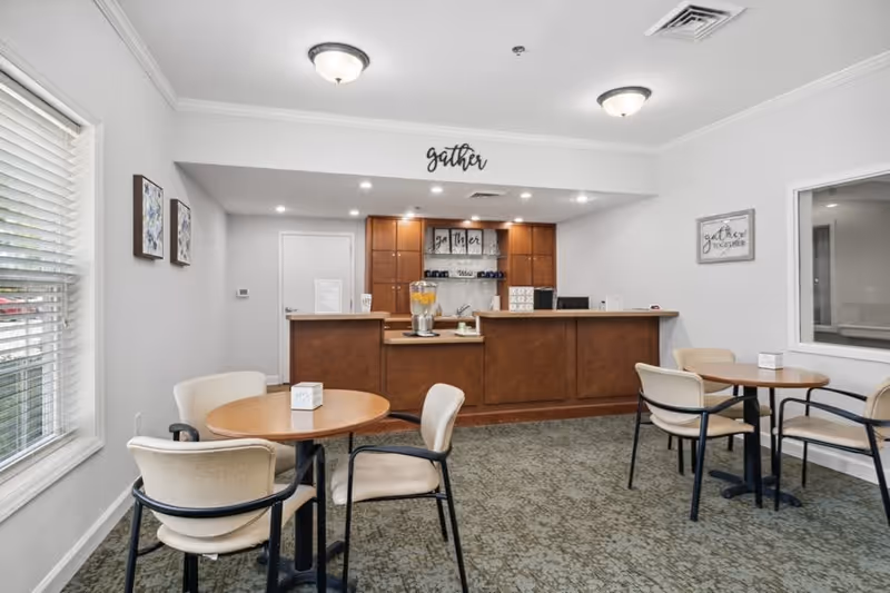 A small dining area with round wooden tables and beige cushioned chairs. In the background, there is a wooden counter with a beverage dispenser filled with lemonade and cups. The walls are light gray with decorative signs that say 'gather' and 'gather together'. There is a window on the right side and two framed pictures on the left wall. The room is well-lit with ceiling lights and natural light from a window on the left.