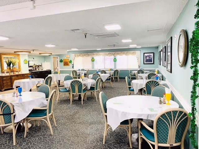 A bright dining room with multiple round tables covered with white tablecloths, each surrounded by green cushioned chairs. The room has light blue walls decorated with framed pictures and a large clock. There are windows at the far end letting in natural light, and some green shamrock decorations are visible, suggesting a festive theme.