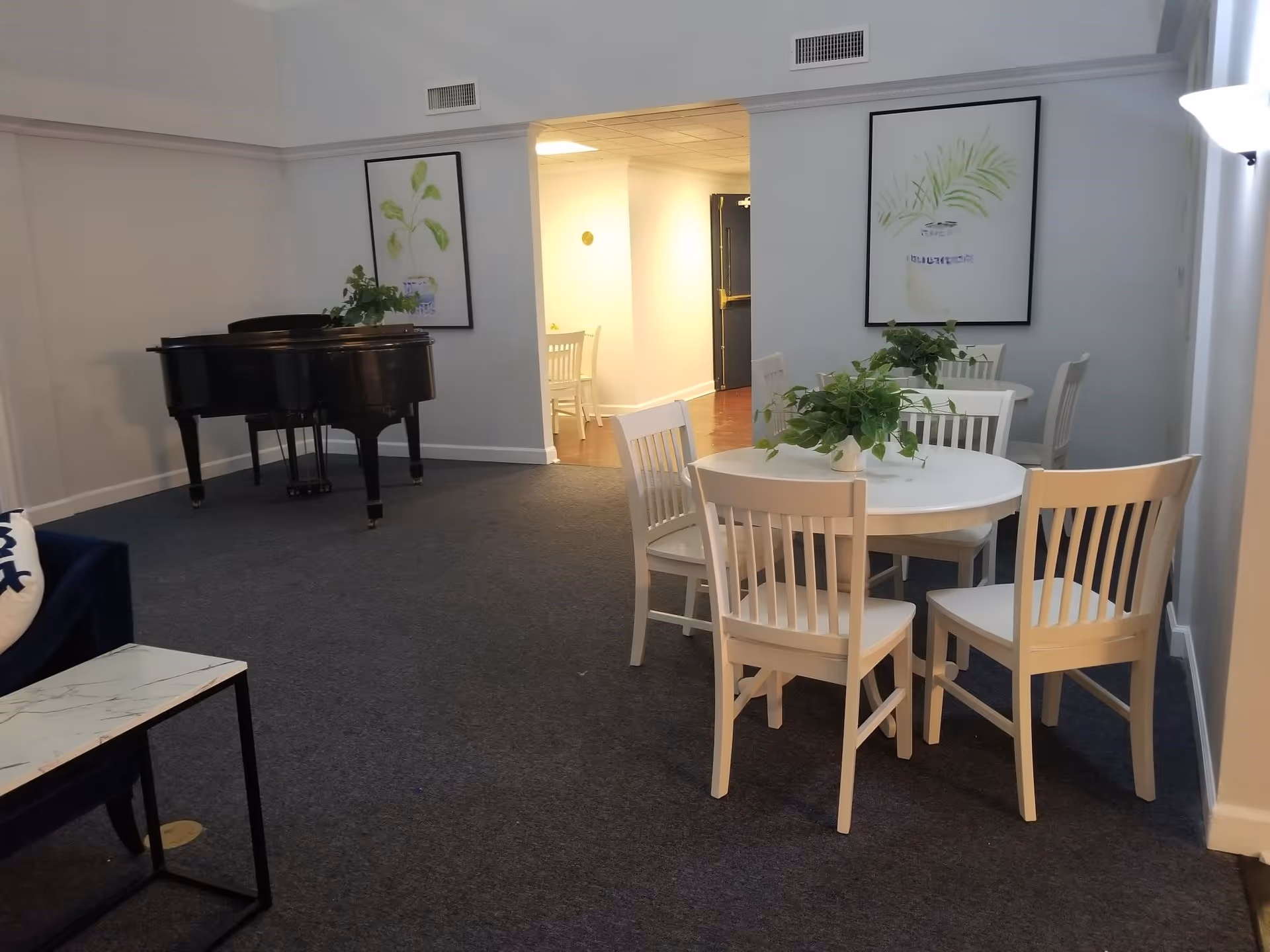 A common area with a round white table surrounded by white chairs, decorated with green plants in the center. In the background, there is a black grand piano and two framed botanical prints on the walls. The room has carpeted flooring and neutral-colored walls.