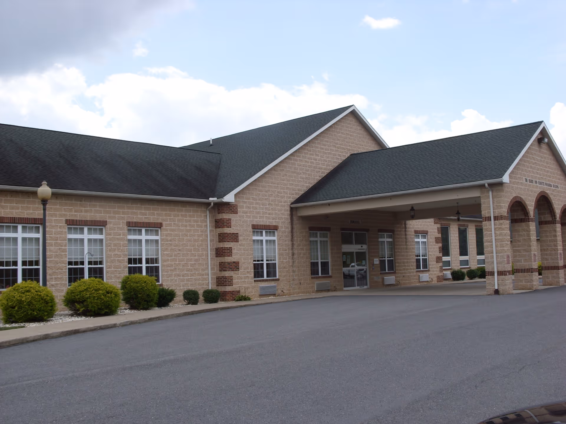 Exterior view of a single-story brick building with a covered entrance and multiple windows, surrounded by small shrubs and a paved driveway under a partly cloudy sky.