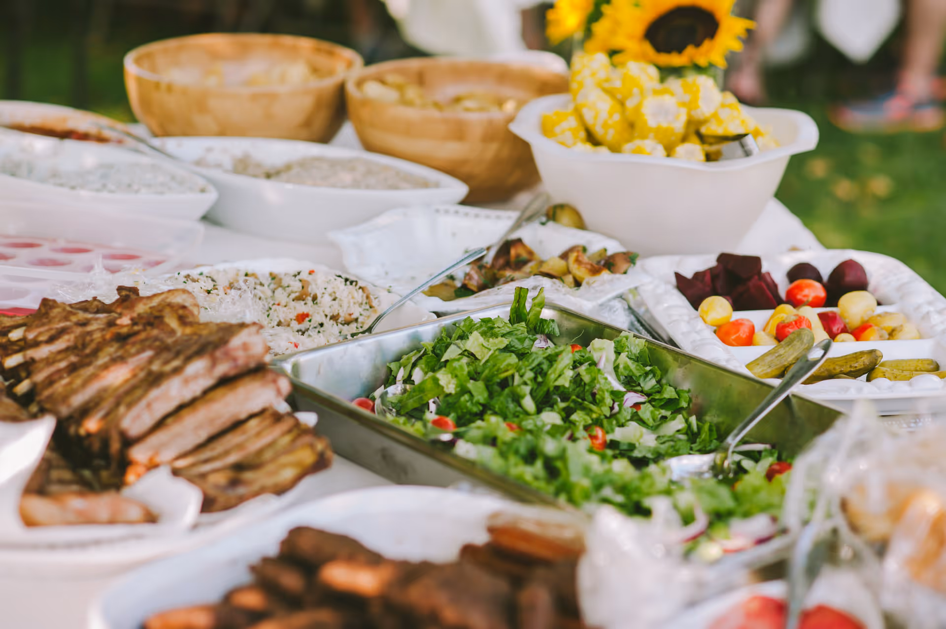 A buffet table with various dishes including grilled ribs, fresh green salad, pickles, corn on the cob, and other side dishes served in bowls and trays outdoors with a blurred background of grass and people.