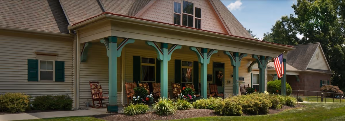 Front exterior view of a senior living facility with a covered porch featuring several rocking chairs, green pillars, and an American flag. The building has beige siding, green shutters, and well-maintained landscaping with bushes and flowers.