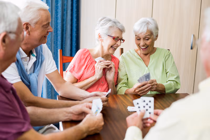 A group of elderly people sitting around a wooden table playing cards, smiling and enjoying each other's company in a bright room with light-colored walls and blue curtains.