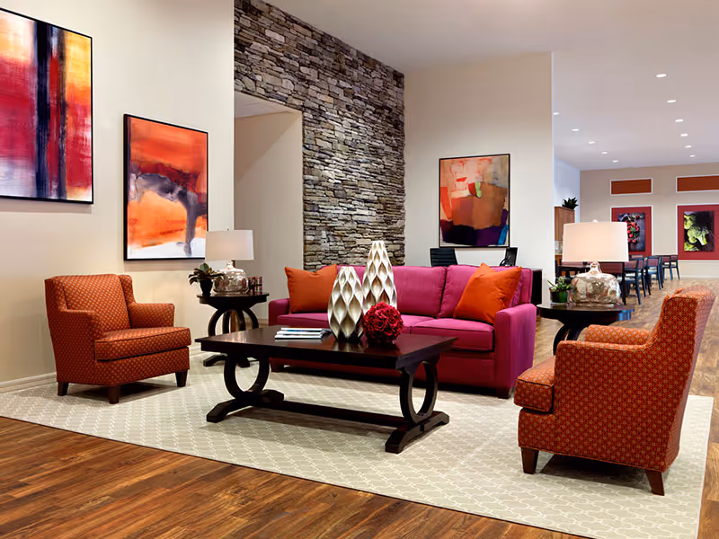 A modern senior living facility lounge area featuring a magenta sofa with orange pillows, two orange patterned armchairs, a dark wooden coffee table with decorative vases and a flower arrangement, side tables with lamps, colorful abstract paintings on the walls, a stone accent wall, and a wooden floor with a light patterned rug.