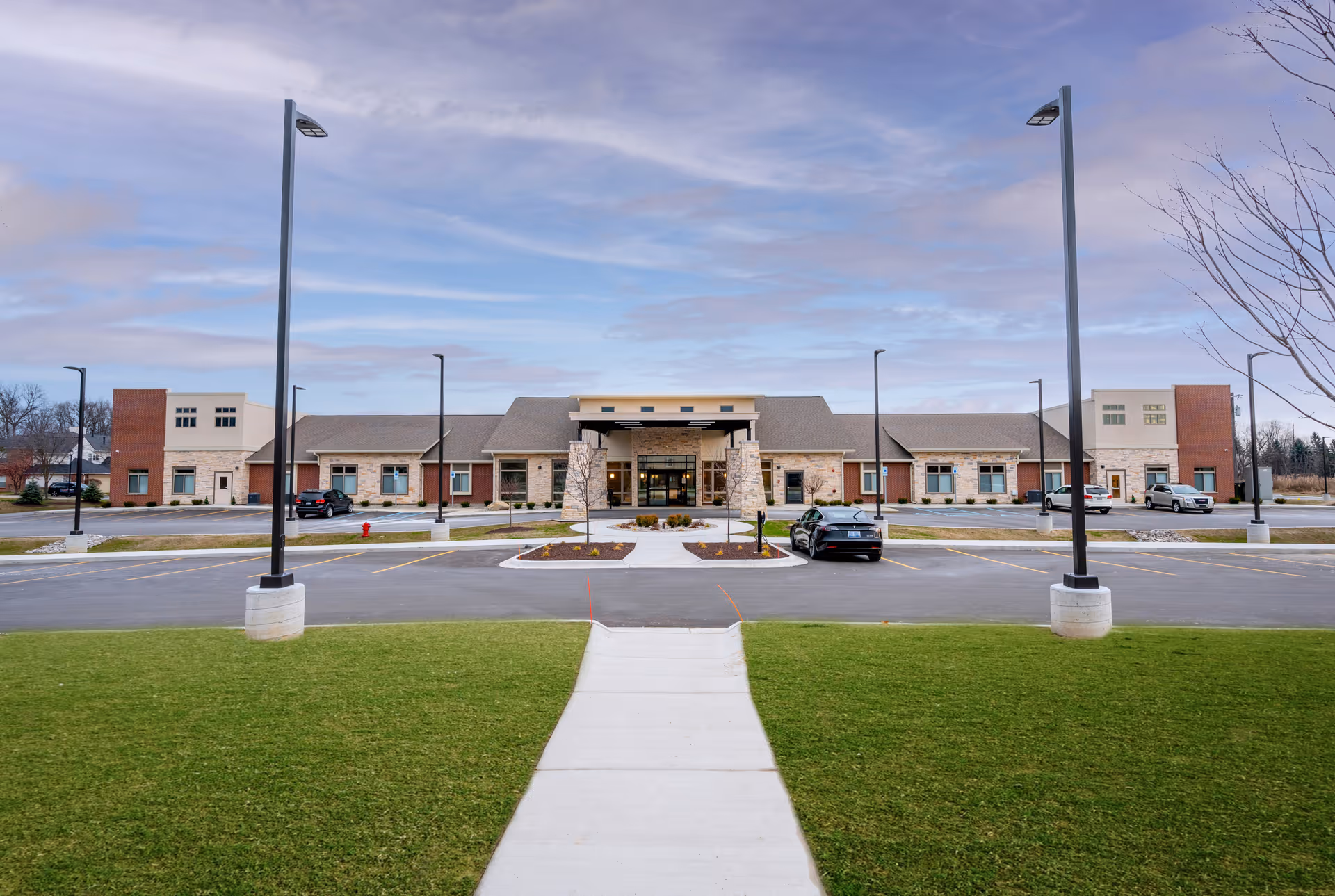 Front exterior view of a single-story senior living facility building with a central entrance, surrounded by a parking lot with several cars and green lawn in the foreground under a partly cloudy sky.
