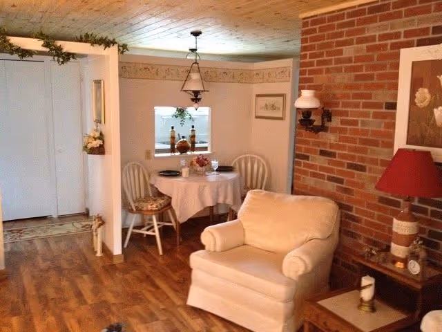 Cozy living area with a beige armchair in front of a brick wall, a small wooden side table with a red lamp and decorative items, and a round dining table covered with a white tablecloth surrounded by two white chairs. The room has wooden flooring and a ceiling light fixture above the dining table. There is a small pass-through window between the dining area and another room.