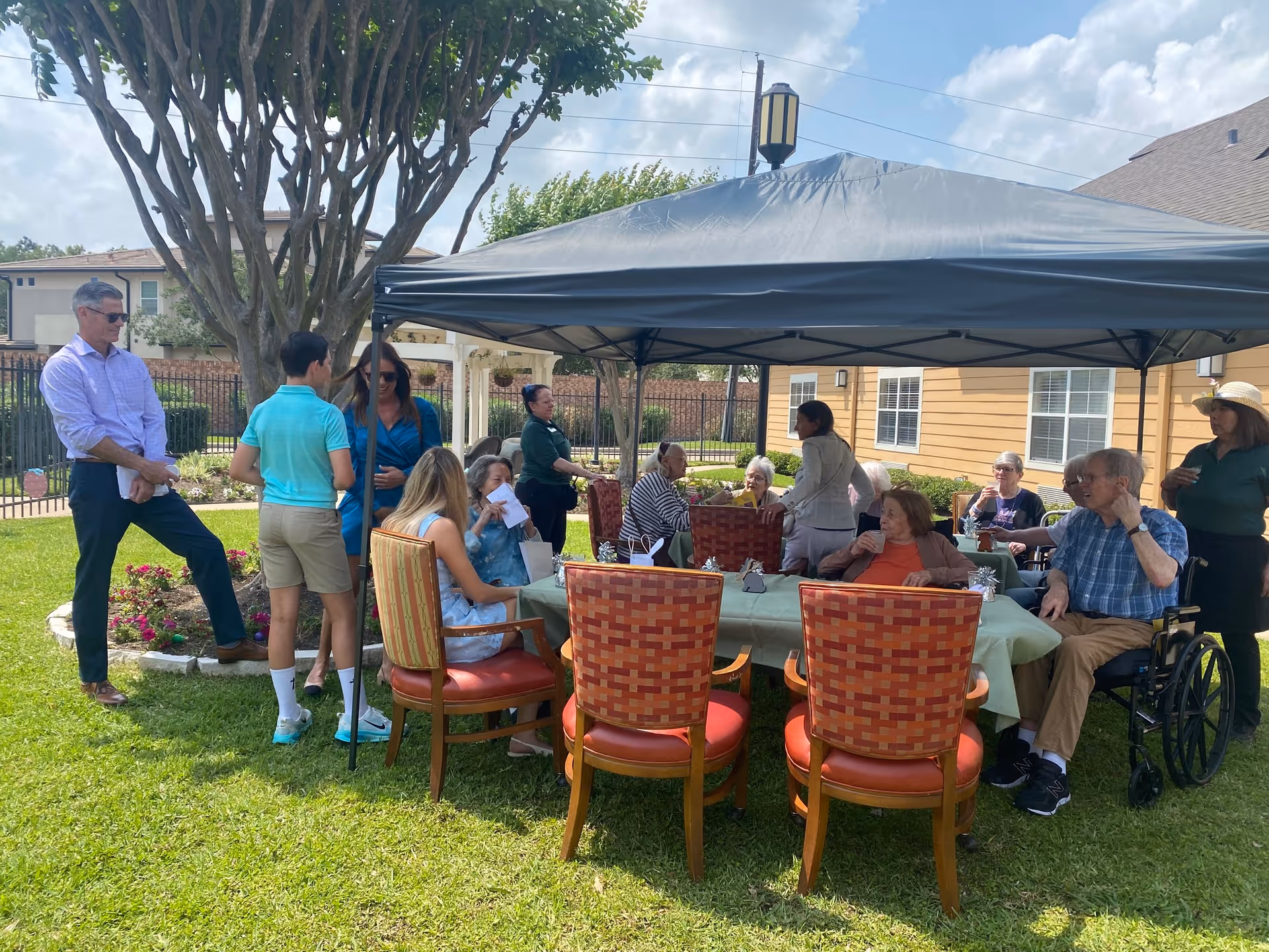 A group of elderly people and caregivers gathered outdoors under a canopy tent in a garden area of a senior living facility. Some are seated around a table with a green tablecloth, while others stand nearby. The setting is sunny with a tree and residential buildings in the background.