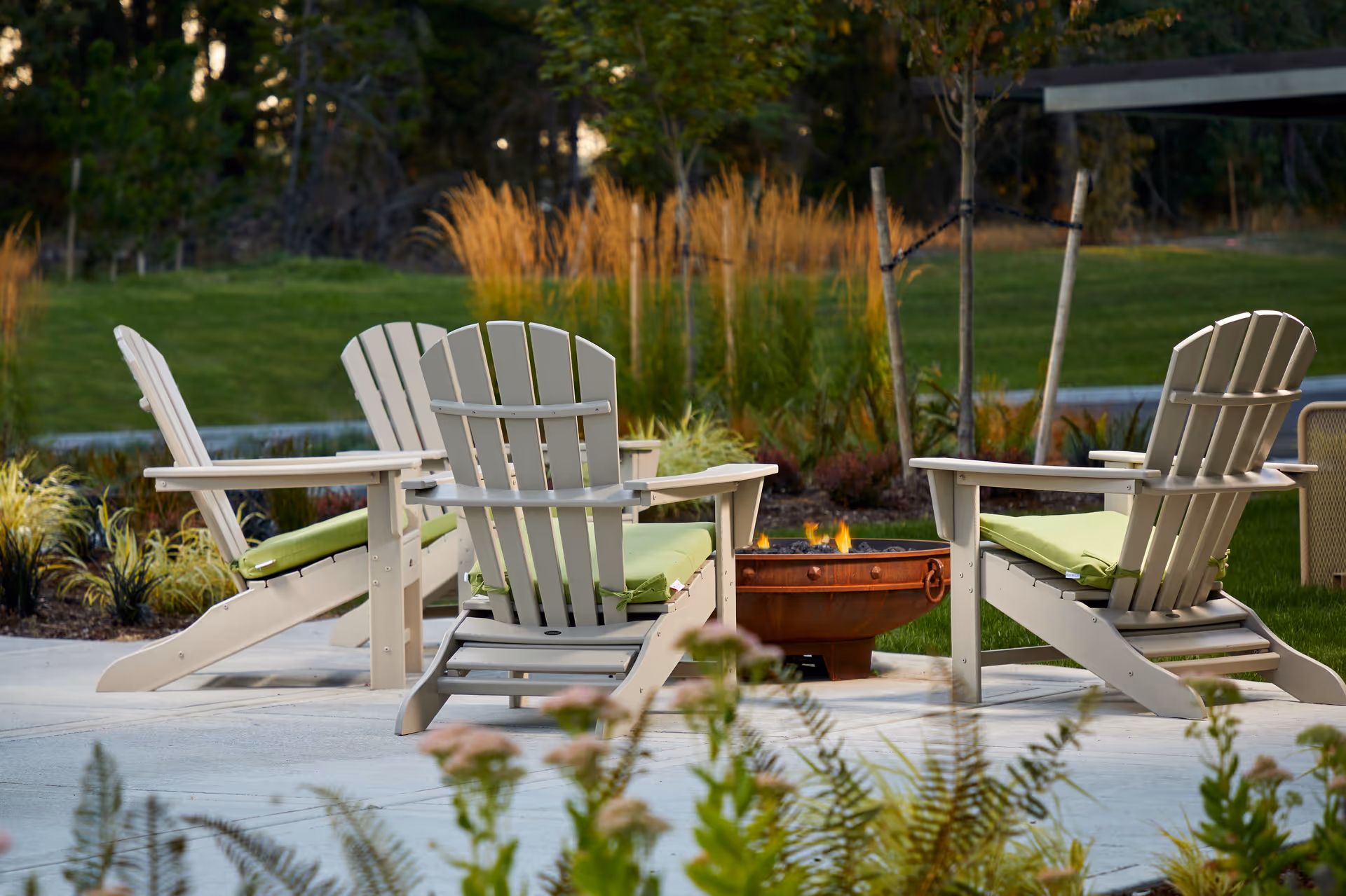Four Adirondack chairs with green cushions arranged around a small fire pit on a patio with lawn and landscaping in the background.