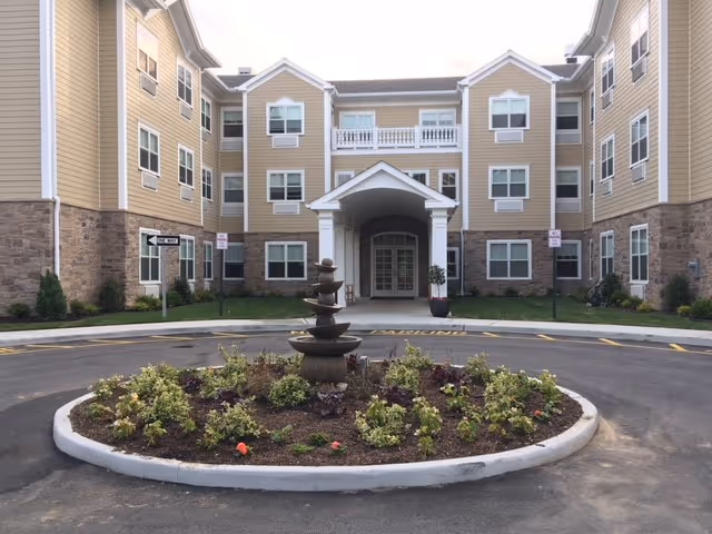 Front exterior view of a three-story senior living facility building with beige siding and stone accents. The entrance features a covered portico with white columns and a balcony above. In front of the entrance is a circular driveway with a landscaped roundabout containing a tiered water fountain and various plants.