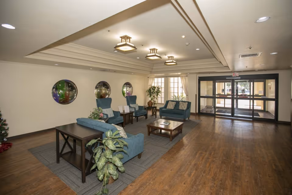 A spacious and well-lit lobby area with wooden flooring, blue upholstered chairs and sofas arranged around wooden coffee tables, potted plants, three circular wall mirrors, large windows with curtains, and glass automatic entrance doors.