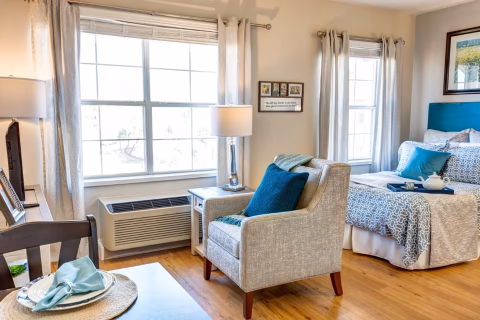 A bright and cozy bedroom in an assisted living facility featuring a bed with patterned bedding and blue accent pillows, a beige armchair with a blue cushion, a side table with a lamp, large windows with curtains, and a small dining table with a place setting.