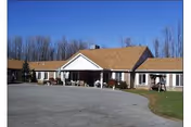 Single-story building with a brown shingled roof and white exterior walls, surrounded by a paved driveway and some grass. The building has a covered entrance with white columns and is set against a backdrop of leafless trees under a clear blue sky.
