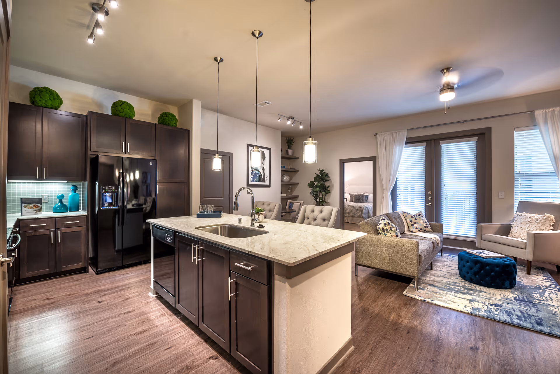 Open-concept kitchen with a marble island and dark cabinets opening to a furnished living area with sofa and armchairs.