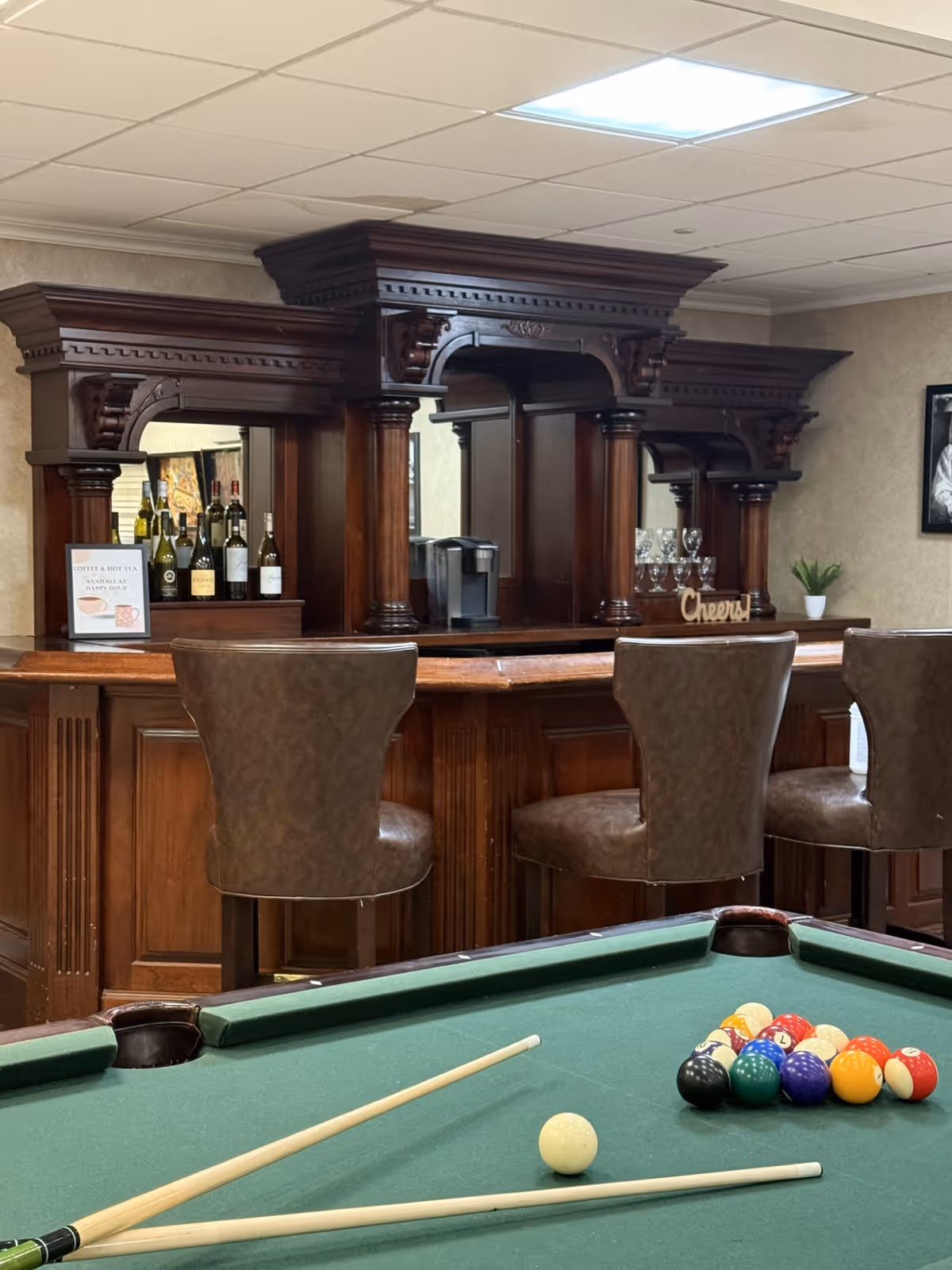 Interior view of a recreational room featuring a green pool table with billiard balls and cues in the foreground. Behind the pool table is a wooden bar with three brown leather bar stools. The bar has an ornate wooden back with shelves holding bottles of wine and glasses. A small sign on the bar reads 'Coffee & Hot Tea Available'.