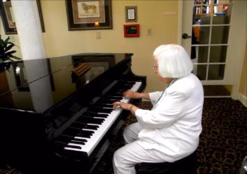 An elderly woman with white hair, dressed in white, is seated and playing a black grand piano in a room with patterned carpet and framed pictures on the wall.