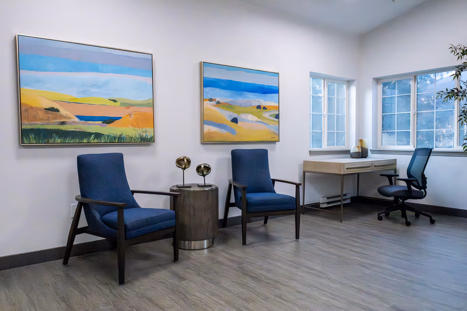 A bright room with two blue armchairs separated by a small round wooden table with two decorative objects on it. On the wall behind the chairs are two colorful landscape paintings. To the right, there is a wooden desk with a black office chair and two windows letting in natural light. The floor has a light wood finish.